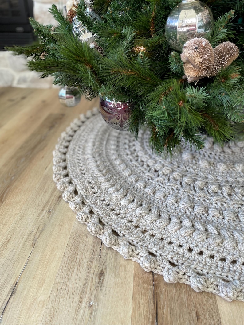 May include: A beige crocheted Christmas tree skirt with a scalloped edge. The skirt is made with a textured stitch and has a subtle sheen.