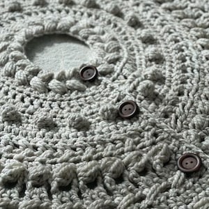 May include: Close-up of a gray crocheted fabric with three brown buttons. The fabric has a textured, bumpy surface.