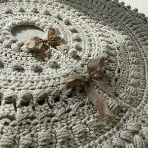 May include: A crocheted tree skirt in a light gray color with a textured design. Two brown satin bows are tied on the skirt.