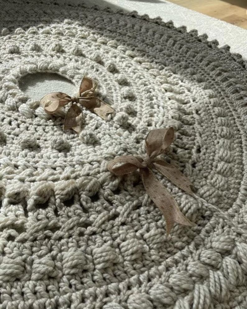 May include: A crocheted tree skirt in a light gray color with a textured design. Two brown satin bows are tied on the skirt.
