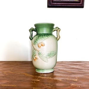 May include: A ceramic vase in light green and cream. The vase has a detailed relief design of leaves and small fruits. It has two handles and a green rim and base. The vase is approximately 25 cm tall.