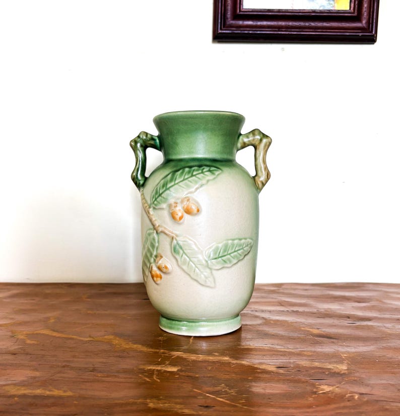 May include: A ceramic vase in light green and cream. The vase has a detailed relief design of leaves and small fruits. It has two handles and a green rim and base. The vase is approximately 25 cm tall.