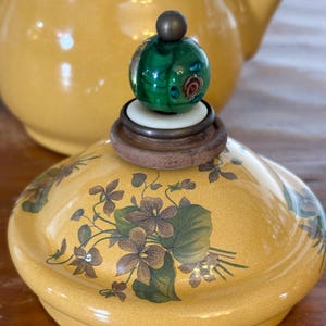Może przedstawiać: Ż&oacute;łta ceramiczna pokrywka do czajnika z kwiatowym wzorem w kolorze brązowym i zielonym. Pokrywka zwieńczona jest zielonym szklanym guzikiem z dekoracyjnym wzorem i małą kulą w kolorze brązu. Czajnik jest w tle.