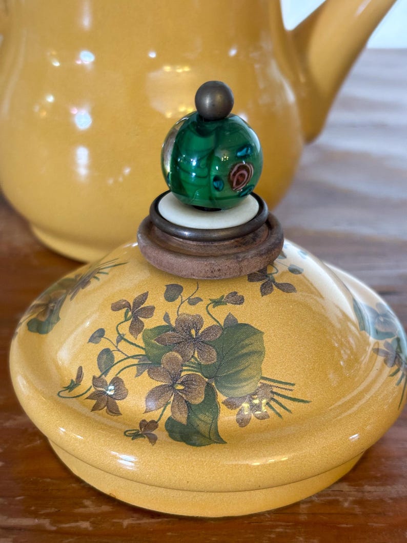 Może przedstawiać: Ż&oacute;łta ceramiczna pokrywka do czajnika z kwiatowym wzorem w kolorze brązowym i zielonym. Pokrywka zwieńczona jest zielonym szklanym guzikiem z dekoracyjnym wzorem i małą kulą w kolorze brązu. Czajnik jest w tle.