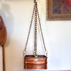 Puede incluir: Una jardinera rectangular de cobre con esquinas redondeadas y detalles acanalados horizontales. La jardinera está suspendida por tres cadenas metálicas que se conectan a puntos de fijación circulares de color latón en el borde superior.