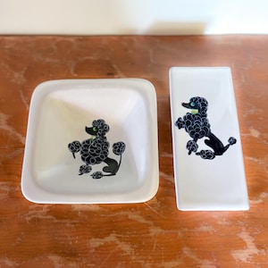 May include: A white ceramic square bowl and a matching rectangular tray, both featuring a black poodle graphic with a green eye and yellow collar. The poodle is depicted in a seated position with curly fur details on both white ceramic pieces.
