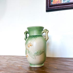May include: A ceramic vase with a light green top and handles, and a beige body. The vase features a relief design of leaves and small orange fruits. The vase is approximately 20 cm tall.