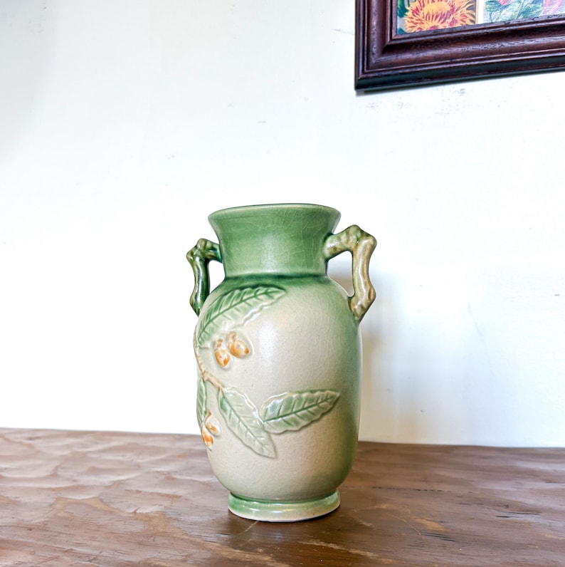 May include: A ceramic vase with a light green top and handles, and a beige body. The vase features a relief design of leaves and small orange fruits. The vase is approximately 20 cm tall.
