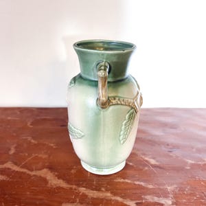 May include: A vintage green ceramic vase with a light green body and a darker green rim. The vase features a handle and leaf-like embossed details. The vase is sitting on a wooden surface.