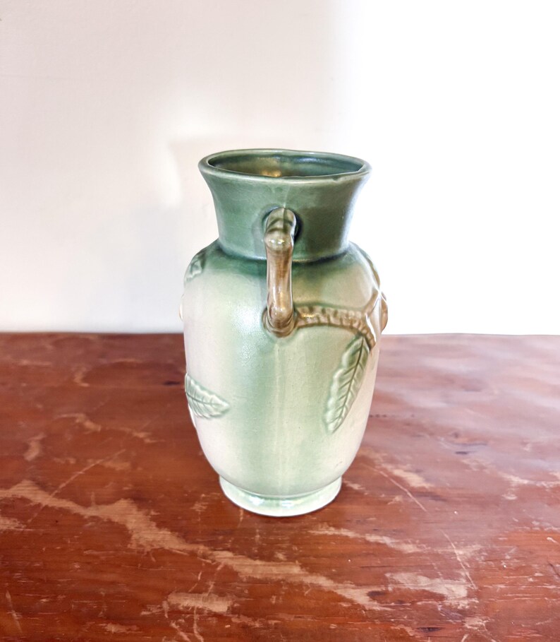 May include: A vintage green ceramic vase with a light green body and a darker green rim. The vase features a handle and leaf-like embossed details. The vase is sitting on a wooden surface.