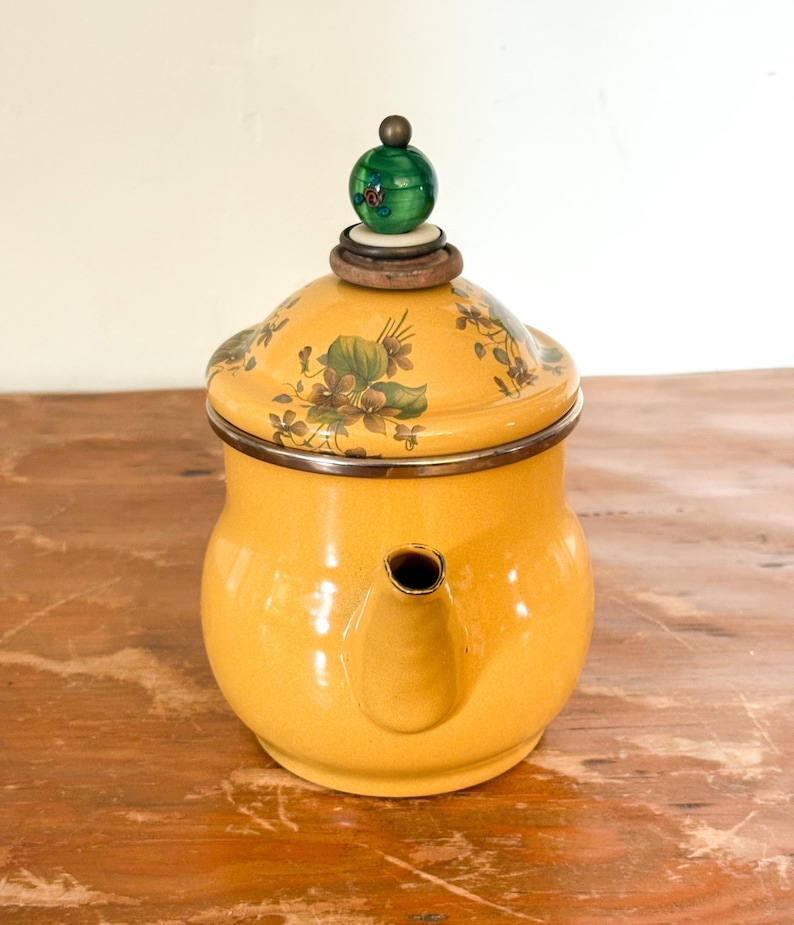 Może przedstawiać: Ż&oacute;łty emaliowany czajnik z kwiatowym wzorem na pokrywce. Czajnik ma srebrną obw&oacute;dkę i zielony uchwyt. Czajnik stoi na drewnianej powierzchni.