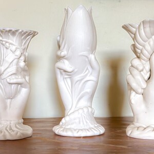 May include: Three white ceramic vases with hand-shaped designs. One vase features a tulip-like form, another a floral arrangement, and the third a classical design. The vases are displayed on a wooden surface.