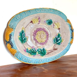 May include: An oval ceramic platter featuring a central purple and yellow chrysanthemum motif surrounded by green leaves. The border is light blue with the embossed text "EAT TO LIVE NOT LIVE TO EAT" and yellow corn-shaped accents on the sides.