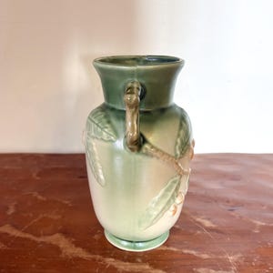 May include: A vintage ceramic vase in shades of green and cream. The vase has a handle and is decorated with a relief design of leaves and berries. The vase has a wide base and a flared rim. The vase is approximately 20 cm tall.