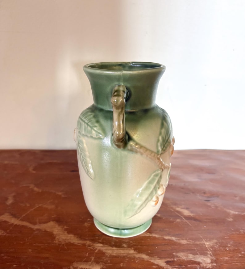 May include: A vintage ceramic vase in shades of green and cream. The vase has a handle and is decorated with a relief design of leaves and berries. The vase has a wide base and a flared rim. The vase is approximately 20 cm tall.
