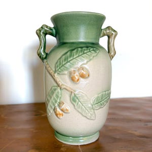 May include: A ceramic vase with a light green rim and handles, and a beige body. The vase features a raised relief design of leaves and acorns in shades of green and tan. The vase is approximately 20 cm tall.