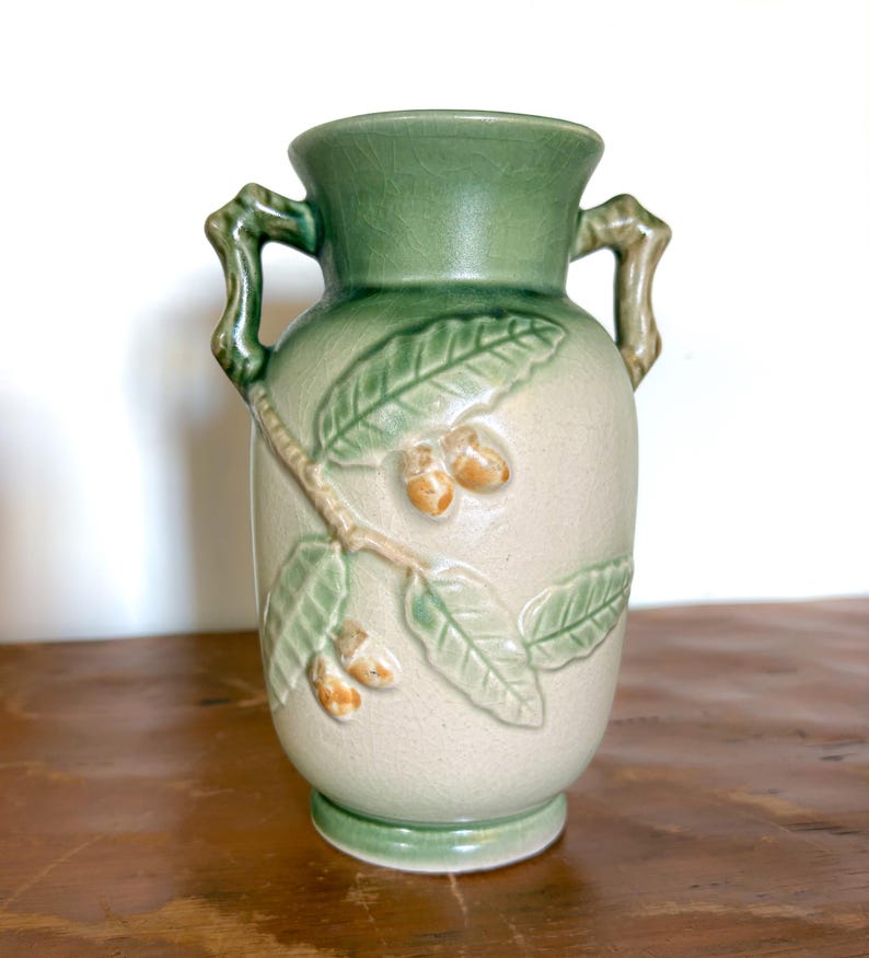 May include: A ceramic vase with a light green rim and handles, and a beige body. The vase features a raised relief design of leaves and acorns in shades of green and tan. The vase is approximately 20 cm tall.