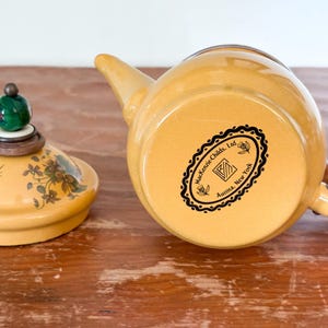 Może przedstawiać: Ż&oacute;łty ceramiczny czajnik z pasującą pokrywką. Pokrywka ma kwiatowe wzory i zielono-brązowe zwieńczenie. Czajnik ma drewniany uchwyt i dzi&oacute;bek. Na spodzie czajnika znajduje się czarno-białe logo z napisem "Mackenzie-Childs, Ltd. Aurora, New York."