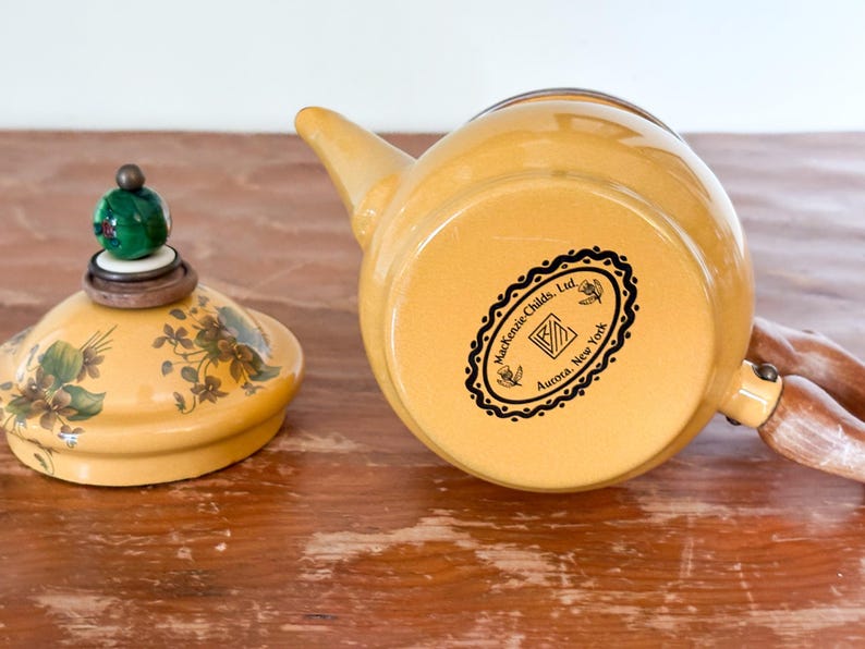 Może przedstawiać: Ż&oacute;łty ceramiczny czajnik z pasującą pokrywką. Pokrywka ma kwiatowe wzory i zielono-brązowe zwieńczenie. Czajnik ma drewniany uchwyt i dzi&oacute;bek. Na spodzie czajnika znajduje się czarno-białe logo z napisem "Mackenzie-Childs, Ltd. Aurora, New York."