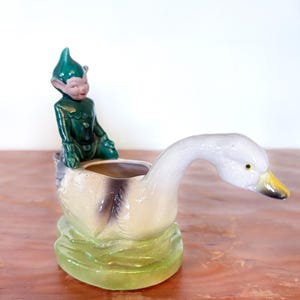 May include: A vintage ceramic planter shaped like a swan, with a green base and a seated elf figure. The swan is white with a yellow beak, and the elf is dressed in green. The planter is likely from the mid-20th century.