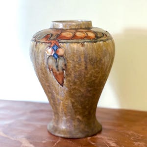 May include: A ceramic vase with a mottled brown and tan glaze, featuring a raised relief pattern around the shoulder. The design includes stylized leaf motifs in shades of orange and brown, accented with small, glossy blue and white diamond shapes.
