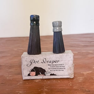 Puede incluir: Dos raspadores de olla negros y verdes con cerdas negras en una caja blanca con el texto "Pot Scraper" y el texto "When you wash dishes if it's pots that you mind this scraper will prove the best aid you can find."