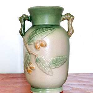 May include: A ceramic vase with a light beige body and green accents. The vase features a design of leaves and acorns, with two handles. The vase is approximately 23 cm tall.