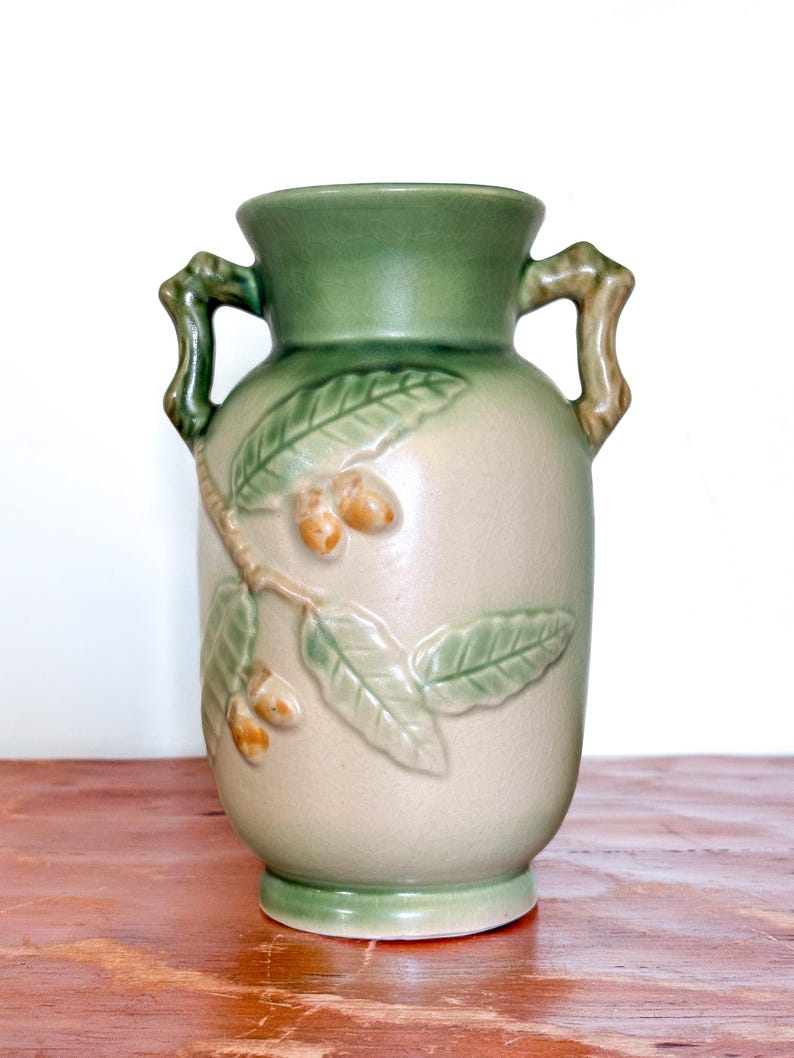 May include: A ceramic vase with a light beige body and green accents. The vase features a design of leaves and acorns, with two handles. The vase is approximately 23 cm tall.