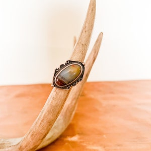 May include: A silver ring with an oval-shaped agate stone set in a decorative frame. The stone is a mix of brown, yellow, and white.