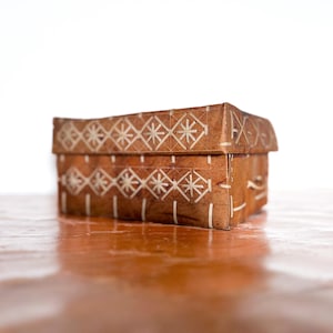 May include: A small, wooden box with a decorative white geometric pattern. The box is made of light brown wood and has a hinged lid.