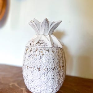 May include: White ceramic pineapple shaped jar with a gold and white pattern. The jar has a lid and is a decorative piece.
