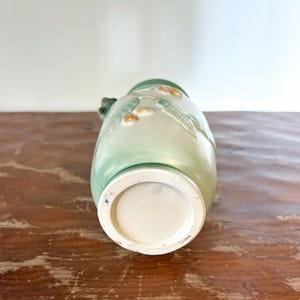 May include: A green and cream ceramic vase with a floral design. The vase has a wide base and a narrow neck. The vase is decorated with raised floral patterns and has a small handle. The vase is sitting on a wooden surface.