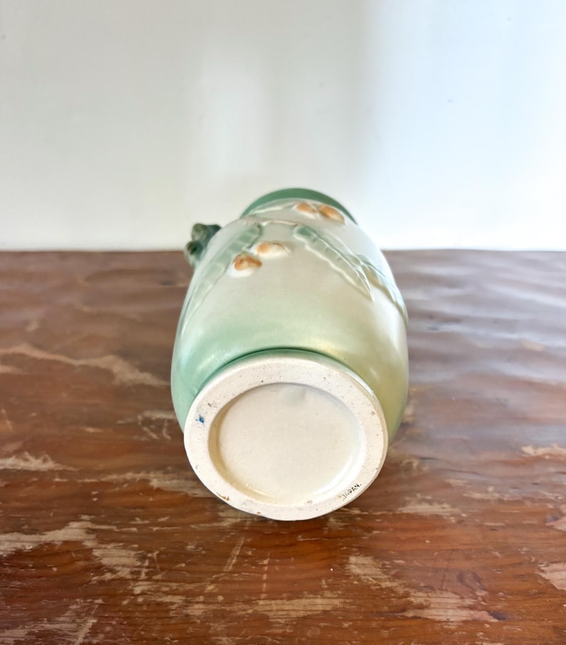 May include: A green and cream ceramic vase with a floral design. The vase has a wide base and a narrow neck. The vase is decorated with raised floral patterns and has a small handle. The vase is sitting on a wooden surface.