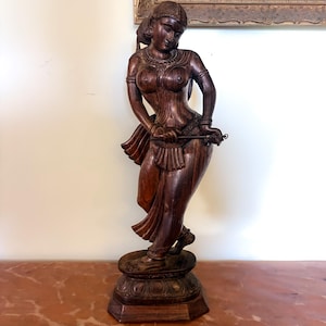 May include: A dark brown carved wooden sculpture of a standing figure in a graceful pose. The figure wears traditional jewelry, including a necklace, armbands, and anklets, and is positioned on a tiered, octagonal base with a lotus petal design.