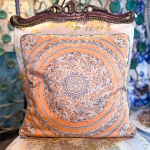 May include: An orange decorative pillow with a detailed floral and leaf pattern. The pillow has a rope trim and is placed on a patterned chair. The design features a central medallion and repeating motifs.