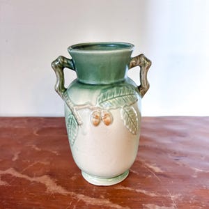 May include: A ceramic vase in shades of green and beige. The top and handles are dark green, the body is a lighter beige with embossed leaf and acorn designs. The vase has a classic urn shape, suitable for displaying flowers or as a decorative item.