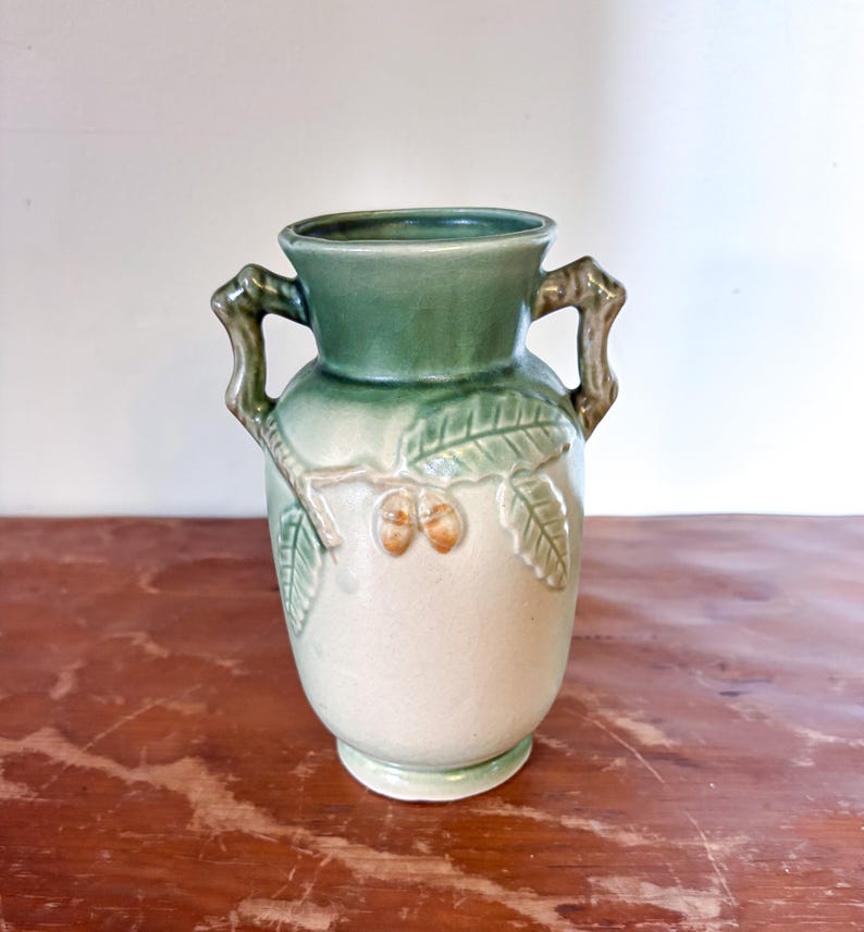 May include: A ceramic vase in shades of green and beige. The top and handles are dark green, the body is a lighter beige with embossed leaf and acorn designs. The vase has a classic urn shape, suitable for displaying flowers or as a decorative item.