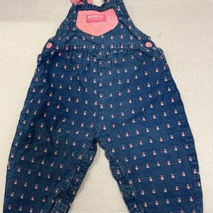 May include: Blue denim baby overalls with pink accents. The overalls feature a pattern of small pink flowers and pink trim on the cuffs and bib. The straps and bib pocket are also pink. The brand name "OshKosh" is visible on the bib.