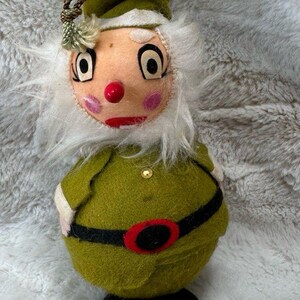 May include: A felt elf figurine featuring a round olive green body, a black belt with a red buckle, and black shoes. The elf has a white faux fur beard, a pointed olive green hat with a brown cord, a red nose, and pink cheeks on a tan face.