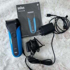 May include: A blue and black Braun Series 3 electric shaver with a wet and dry label, accompanied by a black instruction manual featuring a shaver illustration, a black power adapter, and a black charging cord with a plug.