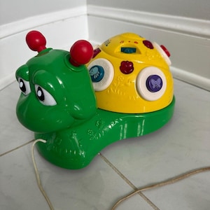 May include: A green plastic pull-along toy shaped like a snail with a yellow shell. The shell features multiple colorful buttons with icons, including a blue button with a spider and a purple button with a butterfly. Two red antennae sit on the head.