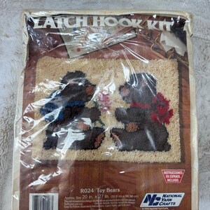 May include: A Latch Hook Kit titled Toy Bears featuring two dark brown bears on a cream background, one with a blue scarf and one with a red bow. The packaging displays the dimensions as 20 in. x 27 in. and includes the National Yarn Crafts logo.