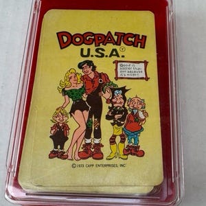 May include: A vintage Dogpatch U.S.A. card set in a clear plastic case with a red base. The cards feature cartoon characters and the text "Dogpatch U.S.A." and "Good is better than evil because it's nicer!!". © 1973 CAPP ENTERPRISES, INC.