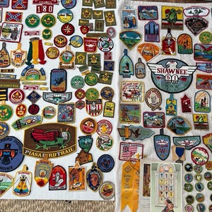 May include: A collection of vintage Boy Scout patches and badges, featuring various shapes, colors, and designs. The patches include text such as "SHILOH", "SHAWNEE", and "THUNDERBIRD DISTRICT". The badges are arranged on a white surface.