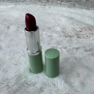 May include: A lipstick with a deep dark red shade and a silver metallic tube base. The cylindrical cap is light green and features the embossed Clinique logo on the top. The lipstick is extended, showing the angled tip of the dark red cosmetic product.