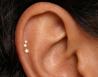 May include: A gold hoop earring with small clear stones set along the outer edge worn in the earlobe, and a gold stud earring featuring three graduated clear stones arranged in a vertical line worn in the upper cartilage of the ear.