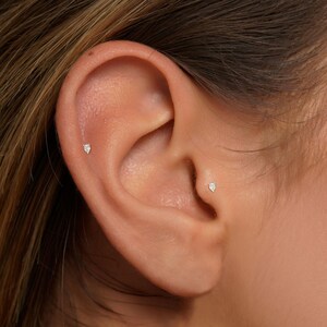 K&ouml;nnte beinhalten: Nahaufnahme eines Ohrs mit zwei kleinen Diamant-Ohrsteckern. Die Ohrringe sind in Gold gefasst und haben eine Tropfenform.