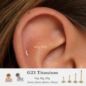 Pode incluir: Um close-up de uma orelha com um pequeno brinco dourado com três pedras preciosas transparentes. O texto "very tiny" está acima da orelha. Abaixo, o texto diz "G23 Titanium" com opções de tamanho em calibre e milímetros.