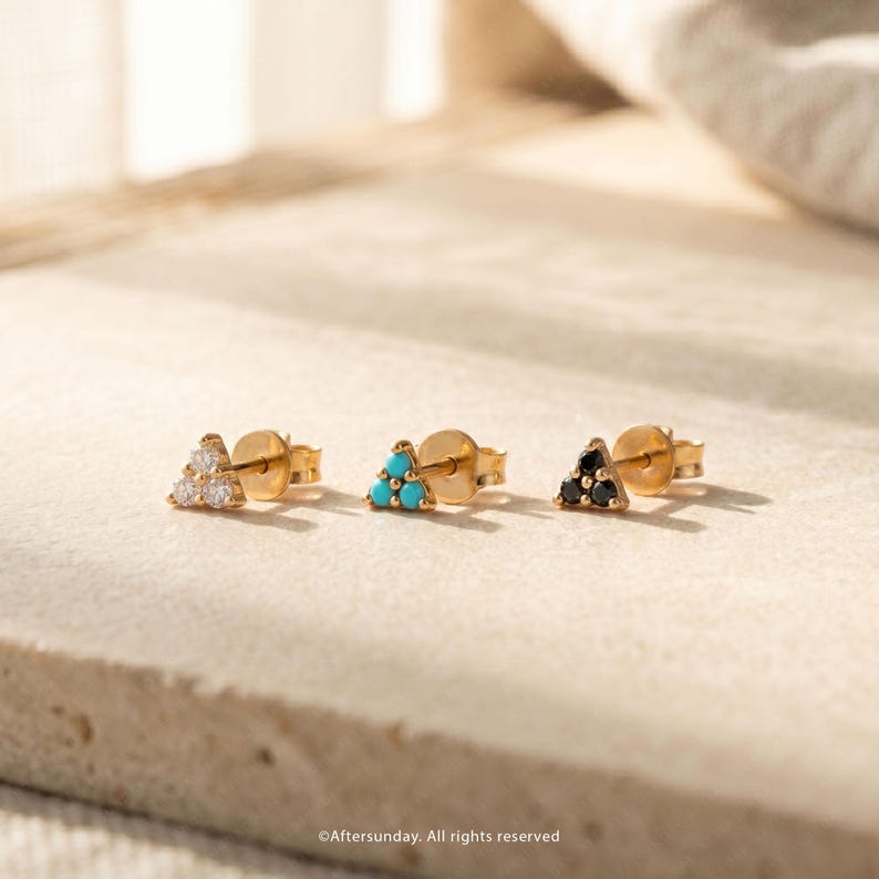 Op de afbeelding: Drie gouden oorknopjes, elk met een driehoekige cluster van drie kleine steentjes. De eerste oorbel heeft heldere witte stenen, de tweede heeft fel turquoise stenen en de derde heeft donkerzwarte stenen, allemaal gezet in een gouden basis.