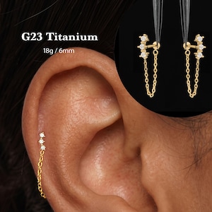 Könnte beinhalten: Ein goldfarbener G23-Titan-Ohrring mit einem vertikalen Stab, drei kleinen klaren Steinen und einer zarten hängenden Kette. Der Text G23 Titanium und 18g / 6mm ist sichtbar. Der Ohrring ist in einem Ohr und in einem kreisförmigen Ausschnitt zu sehen.